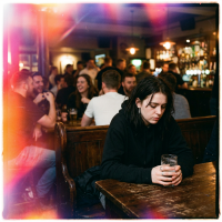 A solitary person sits at a dimly lit bar, holding a glass and looking downcast, while lively groups chat in the background, reflecting the feeling of not wanting to be there.