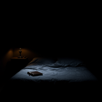 A dimly lit bedroom features a unmade bed with blue sheets. A bedside lamp casts soft light over a closed book resting on the covers, highlighting the contrast between light and shadow.